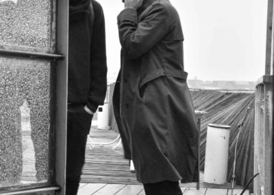 Photo noir et blanc représentant deux hommes sur un toit. Ils sont battus par la pluie et le vent. Artiste, Martes Martes. Lieu, Berlin.