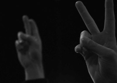 Photo noir et blanc représentant, dans la nuit, une main en l’air montrant le signe “peace“. Artiste, Martes Martes. Lieu, Berlin.