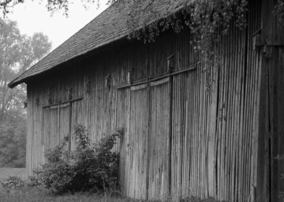 Photo noir et blanc représentant le mur de bois d’une vieille grange. Elle est entourée de végétation. Artiste, Martes Martes. Lieu, Salching.
