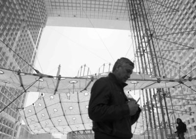 Photo noir et blanc représentant la Grande arche de la Défense derrière un passant. Artiste, Martes Martes. Lieu, Paris.
